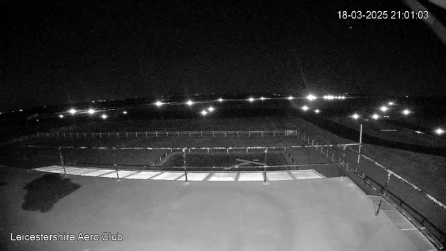 A dark scene captured from a webcam at night, showing a view of an airfield. The foreground features a flat rooftop surface with several lighted areas, possibly reflecting structures beneath the camera. In the background, small lights dot the horizon, indicating distant structures or vehicles in the area. The overall lighting is low, with focused points of brightness contrasting against the dark sky.