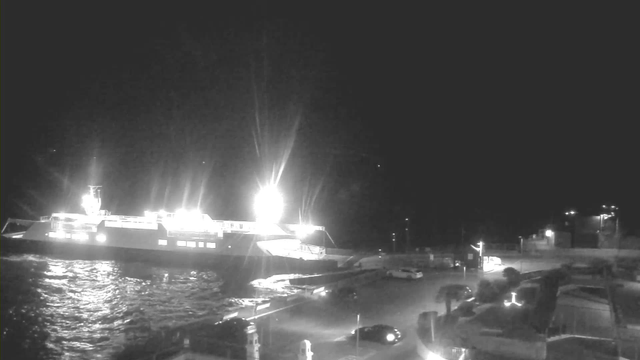 A dark scene showing a large ferry illuminated by bright lights, anchored near a shore. The water reflects the light, creating a shimmering effect. In the foreground, a parking area with a few cars is visible, along with some structures and streetlights in the background. The overall atmosphere is nighttime.