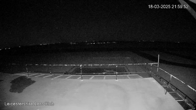 A dimly lit outdoor scene from a webcam showing a flat surface at the forefront, which appears to be a patio or deck area. In the background, a dark sky is visible with a few faint lights indicating distant buildings or features. A faint railing is present, and the area appears empty and quiet. The image captures the nighttime atmosphere with minimal visibility.
