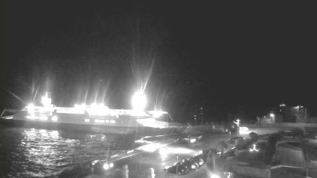 A bright light illuminates a ferry docked at night, casting reflections on the dark water. The ferry is partially visible, with several cars lined up along the dock and some lights from vehicles and nearby structures. The surrounding area is dimly lit, enhancing the contrast between the bright ferry lights and the dark surroundings.