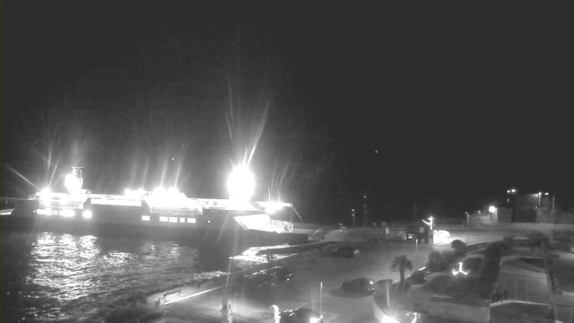A nighttime view of a harbor with a large ferry docked. The ferry is illuminated with bright lights, reflecting on the dark water. There are several parked cars and a pathway leading from the dock area, with additional lights in the background illuminating some structures. The scene is mostly dark with contrasting bright spots from the ferry and street lights.