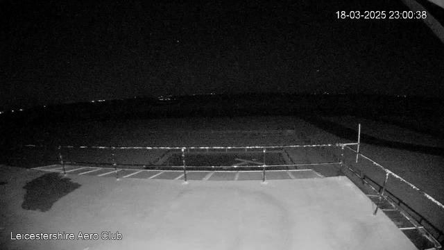 A dimly lit scene captured by a webcam at night, showing a flat rooftop area with a railing on the right side. Below the railing, there are markings on the floor, possibly indicating parking spaces. In the background, faint lights are visible, suggesting distant buildings or other structures beyond the rooftop. The overall atmosphere is dark, with limited visibility of details. The timestamp in the upper right corner reads 18-03-2025 23:00:38.