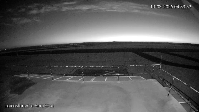 A dimly lit view from a webcam showing a runway or airstrip at dawn. The sky transitions from dark to light near the horizon, with an outline of clouds visible. A barrier or railing is in the foreground, along with landing strips marked on the ground. The scene conveys an early morning atmosphere with no visible aircraft or people.