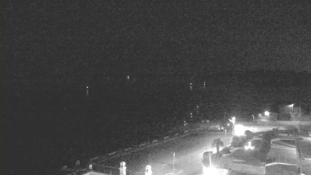 A dimly lit scene captured from a webcam, likely overlooking a waterfront area at night. In the foreground, there are structures and vehicles, with some lights illuminating parts of the scene. The background features a dark body of water, with a few faint reflections visible. The overall atmosphere is calm and quiet, with limited visibility due to the low light conditions.