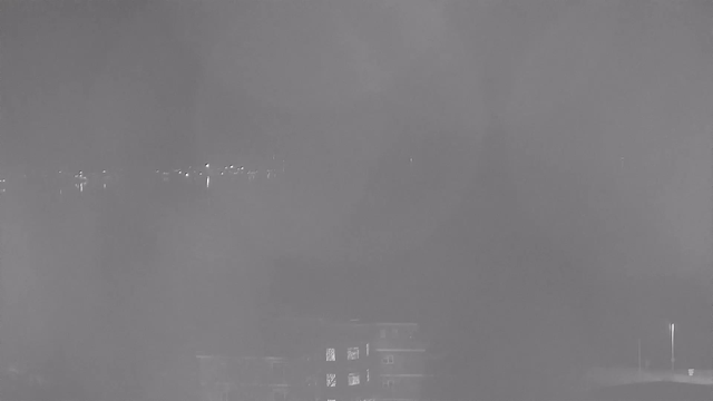 A low-contrast monochrome image featuring a blurry view of a body of water in the background with faint lights reflecting off its surface. In the foreground, there is a dark shape that appears to be a building or structure, with some windows faintly illuminated. The overall atmosphere is hazy and lacks clear details.