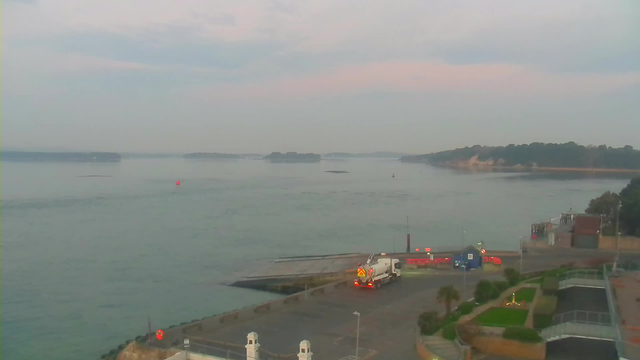 A tranquil view of a harbor at dawn. The water is calm, reflecting soft pastel colors of the sky. In the foreground, a concrete pier extends into the water, and a white truck is parked nearby. There are several small red buoys floating in the water, and in the distance, green islands can be seen. The shoreline features trees and a sandy beach, while a few buildings are visible near the parking area.