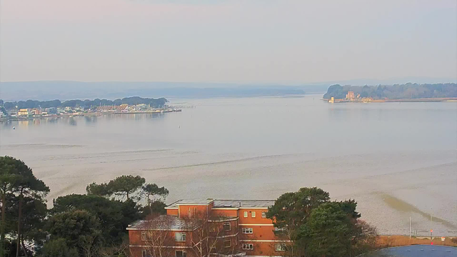 A serene landscape view featuring a calm body of water extending into the distance. On the left, there are several buildings and boats near the shore, suggesting a small harbor. In the background, a gentle slope of trees lines the water, while a distant island is visible on the right with what appears to be a historic structure or tower. The sky has a soft, light hue, indicating a peaceful, early morning or late afternoon. The foreground includes greenery and a few residential buildings close to the water's edge.