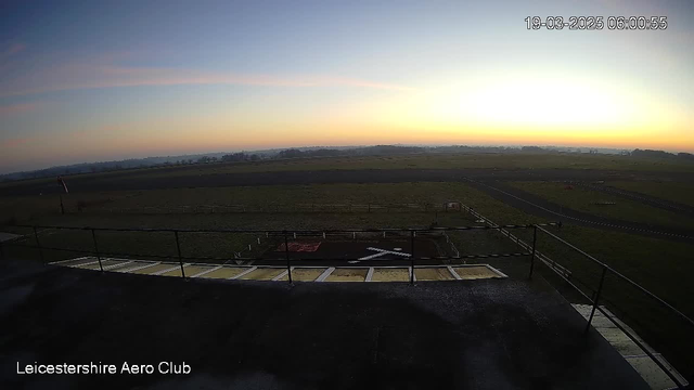 A wide view of a clear sky at dawn, transitioning from light blue to soft orange and pink hues near the horizon. In the foreground, there is a rooftop railing and a light-colored surface leading to an area marked with red and white, resembling a landing or takeoff zone. In the distance, lush green fields stretch out, with a few scattered trees and what appears to be a runway or road. The scene captures a peaceful, early morning atmosphere at an aerodrome.