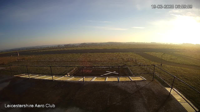 A view from a webcam at the Leicestershire Aero Club, featuring a wide, open landscape under a clear blue sky with faint wispy clouds. In the foreground, there is a grassy area with a white "X" marking on the ground, indicating a helipad or landing area. A railing surrounds the area, and in the background, rolling hills can be seen, fading into the horizon. The lighting suggests that it is early morning.