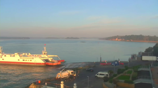 A serene waterfront scene during early morning light. In the foreground, a large white and orange ferry is docked at the terminal, with its ramp extended towards the land. Nearby, a white vehicle is parked on the pavement, with a road sweeper truck visible. The water is calm and reflects the soft pastel colors of the sky, which transitions from light blue to hints of orange and yellow near the horizon. In the background, a wooded area and cliffs can be seen along the shoreline, and small boats are visible further out in the water.