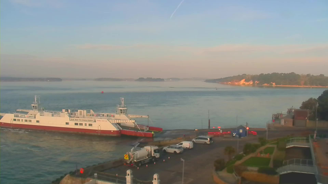 A ferry with a white hull and red lower section is docked at a harbor, facing the water. The background shows calm water with a gentle reflection of the sky, which is clear with some light clouds. In the distance, there are small islands and a tree-lined shore. Several vehicles are parked nearby, including vans and cars. The scene is bathed in soft morning light, creating a serene atmosphere.