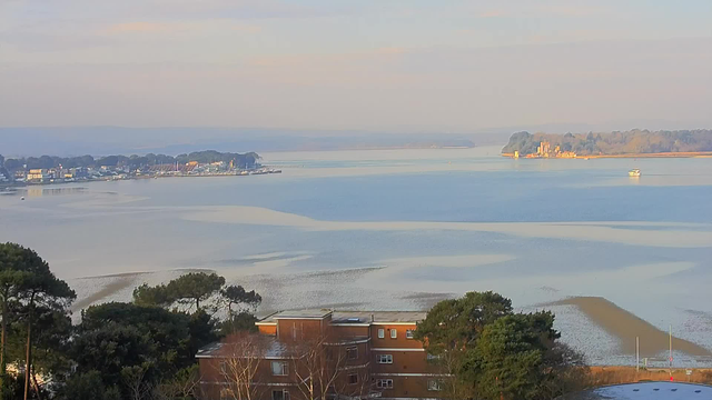 A scenic view of a calm body of water, with a sandy shore in the foreground and gentle waves lapping at it. To the left, a small marina with several boats docked. The opposite side features an island with trees and a structure that appears to be a castle or fort. The sky is light blue with soft clouds, suggesting a peaceful morning atmosphere.
