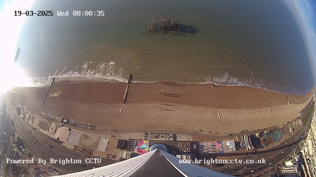 Aerial view of a beach with golden sand and gentle waves lapping at the shore. A partially submerged pier can be seen in the water, with some structures visible in the background. The coastline features a promenade with a few people walking. Bright, sunny weather creates a sparkly effect on the water's surface. The scene is viewed from a high angle, with a modern building at the bottom of the image. The date and time are displayed at the top left corner.