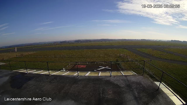 A clear blue sky with wispy clouds is visible above a wide, grassy field. In the foreground, there is a railing enclosing a small platform. Below the platform, a landing area marked with a white cross is present on a dark surface. In the distance, an airstrip extends across the horizon, bordered by more fields. A few trees are faintly visible far away, under a bright and sunny atmosphere. The time and date display in the top right corner shows "19-03-2025 08:59:56."