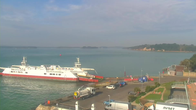A view of a calm harbor with a large white and red ferry docked at the terminal on the left. The water is turquoise with a few small boats in the distance. To the right, there are parked vehicles, including a grey truck and several cars, as well as green grass and a pathway leading to a small building. In the background, there are trees and a rocky shoreline under a blue sky with some clouds.