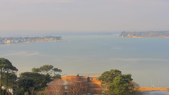 A serene view of a large body of water, bordered by a coastline with boats docked at a marina. The scene includes soft, hazy colors, with gently rolling hills in the background. In the foreground, there are trees and an orange building, likely a residential structure, partially obscured by greenery. The sky above is pale and appears overcast, contributing to the tranquil atmosphere of the landscape.