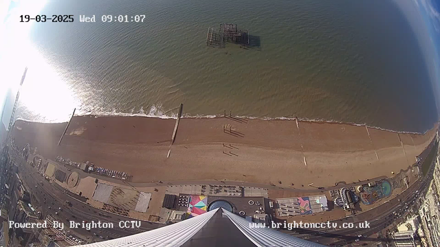A high-angle view of a coastline showing a sandy beach and the sea. The beach extends horizontally across the image, with a pier jutting out into the water. In the distance, remnants of a structure can be seen partially submerged in the sea. Below, there are urban elements including a colorful patterned area and some buildings. The image is bright, reflecting sunlight on the water's surface. A timestamp indicates the date and time as 19th March 2025, 09:01:07. The text "Powered by Brighton CCTV" is displayed at the bottom.