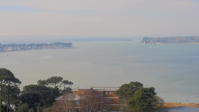 A panoramic view of a calm body of water with a subtle grayish-blue hue. In the distance, there are small boats and a marina with several masts visible. The shoreline on the left features buildings and trees, while the right side shows a more wooded area and a distant landmass. The sky above is light and hazy, suggesting a soft, overcast day.