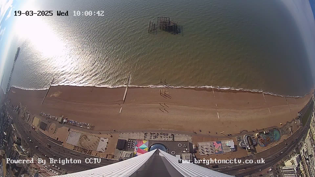 Aerial view of a sandy beach with gentle waves lapping at the shore. The coastline features several structures, including a pier extending into the water and remnants of an old pier slightly submerged. Below the beach, a colorful boardwalk area displays various patterns. The sky above is bright with a few clouds, and the sun reflects off the water's surface, creating a shimmering effect. The timestamp in the upper corner indicates the date and time: 19th March 2025, 10:00:42. There is text at the bottom indicating "Powered by Brighton CCTV."
