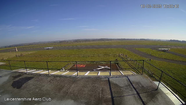 A wide view of an airfield on a clear day. The sky is bright blue with a few wispy clouds. In the foreground, there is a railing indicating a viewing area. Below, a grassy area features a marked helipad with an "X" pattern. In the background, a small airplane is parked on the runway, and the landscape extends into the distance with rolling hills. A few fences outline the airfield. The time stamp indicates the image was captured at ten o'clock.