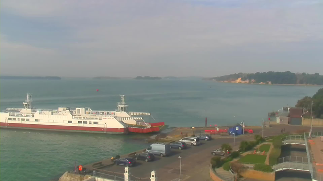 A ferry boat is docked at a harbor with a red and white color scheme. The boat is moored near a parking area filled with cars. In the background, calm blue waters extend towards the horizon, where there are several small islands visible. A nearby shoreline features trees and sandy hills, while the sky is partly cloudy with a soft light.