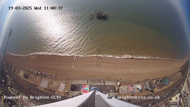 A panoramic view from a high vantage point overlooking a beach. The sandy shoreline stretches across the bottom of the image, with gentle waves lapping at the shore. A few people are walking along the beach. In the distance, there is a pier extending into the water, and an old, partially submerged structure can be seen in the ocean. The beach area features colorful beach huts and amusement rides, with a vibrant pattern on the ground. The scene is bright and sunny, with a clear sky reflecting off the water.