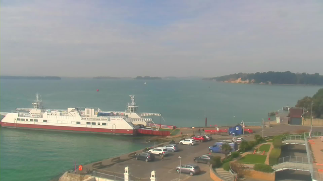 A large white ferry with red accents is docked at a pier on a calm body of water. The shoreline is visible in the background with green trees and rocky formations. Various vehicles are parked near the water, including cars in a parking lot. The sky is light blue with scattered clouds, creating a serene atmosphere.