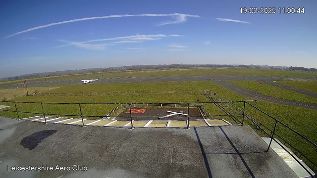 A clear blue sky with a few white clouds overhead. In the foreground, a small white aircraft is positioned on a runway. The grassy area surrounding the runway is visible, along with some wooden fencing. To the left, there is a marked helipad featuring a white cross. The scene is tranquil with no visible aircraft taking off or landing.