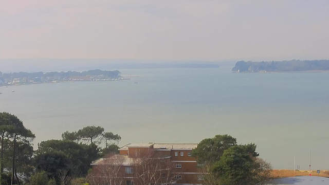 A tranquil view of a calm body of water extending into the distance, with a hazy sky above. On the left side, there is a marina with several boats and yachts docked. In the foreground, there are patches of greenery including trees, and a brick building with multiple windows. The shoreline on the right features a series of trees and possibly a distant structure, adding to the scenic coastal atmosphere.