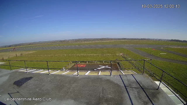A clear blue sky overhead with a few wispy clouds. In the foreground, there is a flat rooftop with a railing, and below it, a grassy area leads to a runway. A helipad marked with a white cross is near the bottom left, and a red sign is visible on the right side of the image. The grassy area is bordered by a low wooden fence. The runway stretches into the distance, with markings seen on the asphalt.