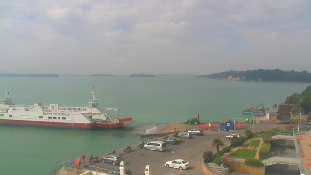 A view of a harbor scene with calm, turquoise waters. A large ferry boat, white with red trim, is docked at the pier. Several vehicles are parked nearby on a paved area, and there are a few people visible, including some in work attire. In the background, green hills and several small islands are visible in the distance, under a partly cloudy sky.