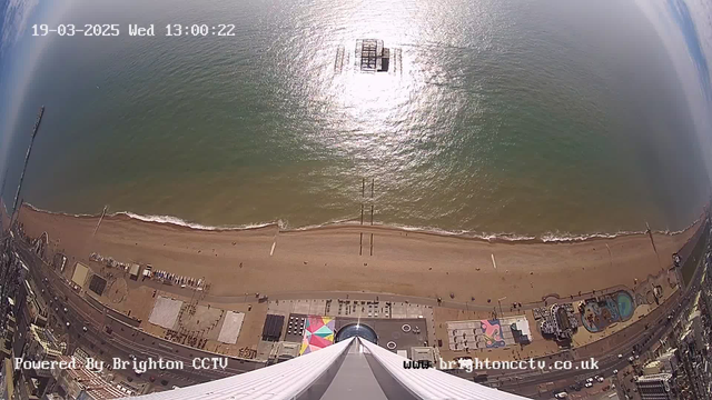 A panoramic view from a high vantage point above a coastline. The beach stretches below with a sandy area, where a few people can be seen walking. There are scattered colored beach umbrellas and sunbathers on the sand. The ocean is visible, reflecting sunlight and showing gentle waves. A distant pier structure extends into the water, and several small boats are noticeable on the surface. The left side of the image shows parts of a cityscape with roads and buildings, while the sky is partly cloudy. A timestamp indicates the date and time as 19th March 2025, 1:00 PM.