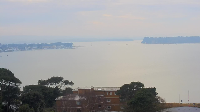 A panoramic view of a calm body of water under a partially cloudy sky. In the distance, there is a shoreline with trees and buildings. Small boats can be seen on the water, and the overall atmosphere is serene, with soft colors and gentle light reflecting on the surface. The foreground features some greenery and a building with several windows.