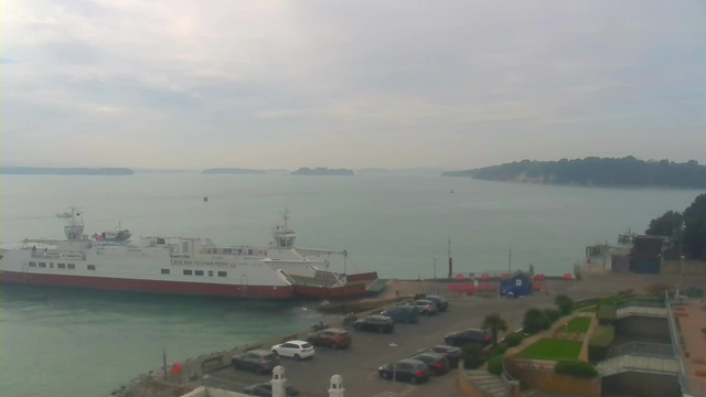 A view of a calm sea with a large white and red ferry docked at a pier on the left side. Several cars are parked in a lot nearby. In the background, there are small islands on the horizon and a coastline with trees, under a cloudy sky. The water appears still and reflects the overcast light.