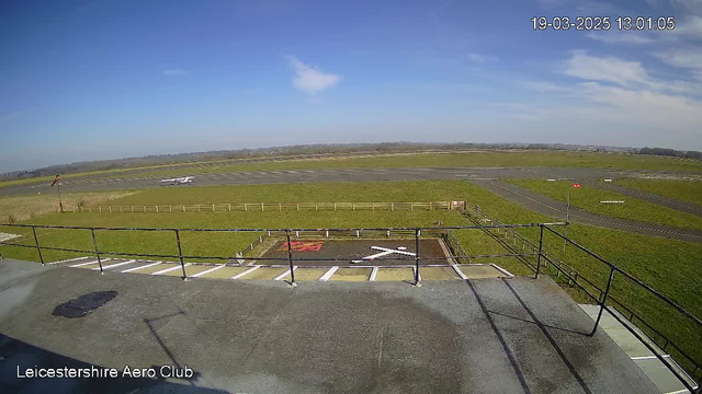 A view from a high vantage point at an airfield. The foreground features a railing, and below it is a helipad marked with a large white "X" on a grassy area. The background shows a wide expanse of grassy land and an asphalt runway, with light traffic markers visible. The sky is mostly clear with a few clouds, and the scene is bright due to daylight. The overall setting suggests a tranquil day at the Leicestershire Aero Club.