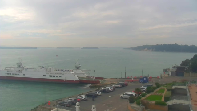 A ferry with a white body and red lower section is docked at a harbor. The water is calm and greenish-blue, with a few small boats in the distance. In the foreground, there is a parking lot with several cars and some greenery, including flower beds and steps leading down. The sky is overcast with gray clouds, hinting at a cloudy day. On the coastline in the background, there are trees lining the shore.