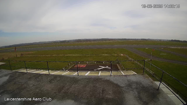 A view from a webcam located at Leicestershire Aero Club, showcasing a wide-open grassy area adjacent to an airstrip. The scene features a cloudy sky, a flat green field, and a section of the runway with markings. In the foreground, there is a railing visible along the edge of a building, and a large white "X" is painted on the ground below, indicating a landing or takeoff area. The landscape extends further back, with a distant horizon.