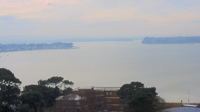 A serene view of a calm body of water under a pale sky. In the foreground, there is a building surrounded by trees. The midground features a shoreline with several boats and a distant landmass lined with trees. The background fades into a hazy horizon, where the water meets the sky. Overall, the scene conveys a peaceful, tranquil atmosphere.
