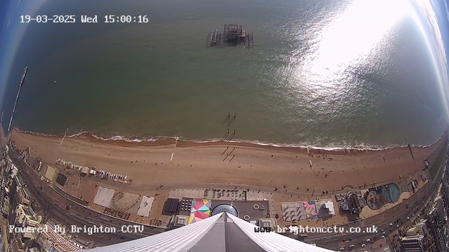 A view from a high vantage point overlooking a sandy beach and the ocean. The sun glimmers on the water, creating a bright reflection. In the distance, a derelict pier stands partially above the water. Along the beach, small figures of people can be seen walking, with some shadows cast on the sand. At the bottom of the image, colorful structures and attractions along the waterfront are visible, including a circular building and a swimming pool area. The sky is mostly clear with some clouds.