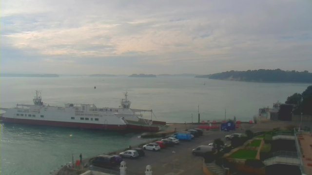 A view of a calm harbor with a large white and red ferry docked at the pier. In the foreground, several cars are parked along the waterfront, with some greenery and pathways visible. The background features a misty skyline with distant islands and a cloudy sky, adding a tranquil atmosphere to the scene.