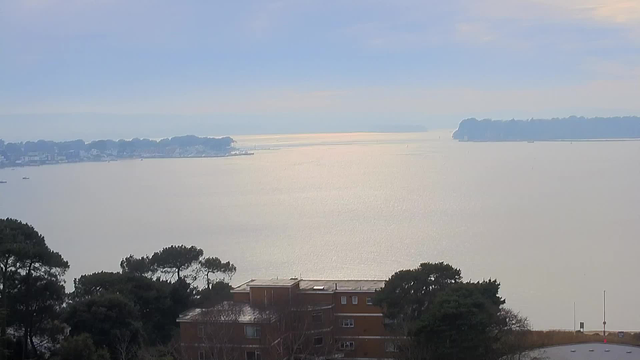 A calm water landscape is visible with a luminous reflection from the sky creating a silvery sheen on the water's surface. In the distance, a shoreline is framed by trees and buildings, suggesting a coastal scene. The sky is mostly cloudy with soft blue and gray hues, indicating overcast weather. Near the bottom of the image, there are dark green trees and a brown building, adding depth to the view.