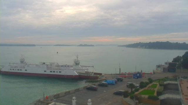 A view of a calm harbor under a cloudy sky. A large white ferry with red accents is docked at the port. In the background, several small boats are visible, and green hills can be seen along the shoreline. The water is a soft blue-green. At the dock, there are parked cars and some construction or maintenance equipment. The atmosphere is quiet and serene.