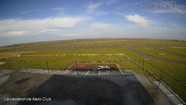 A clear view of a grassy airfield under a bright blue sky with scattered clouds. In the foreground, a fenced area surrounds a marked landing zone with a red and white "X" symbol on a dark surface. The runway is visible, leading into the distance, with a white runway edge line and a red windsock indicating wind direction. In the background, fields extend towards rolling hills. The scene is bathed in natural daylight, conveying a calm and open atmosphere.