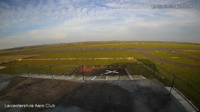 A wide view of an airfield showing a grassy landing strip and a few runways in the background. There are white markings on the ground resembling an airplane takeoff position. A fence runs along the edge of the field, and a wind direction indicator is visible on a pole to the left. The sky is mostly clear with some clouds. The image is taken during the late afternoon.