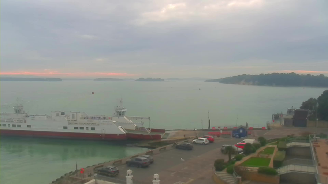 A gray, overcast sky hangs over a tranquil harbor. In the foreground, a large ferry boat with a white and red color scheme is docked near the shore, where some vehicles are parked. The harbor water appears calm and reflective, with hints of pink on the horizon suggesting dawn or dusk. In the background, lush, green hills outline the shoreline, while a few small boats are visible in the water. There is a small building at the edge of the harbor and some landscaped areas with palm trees nearby.