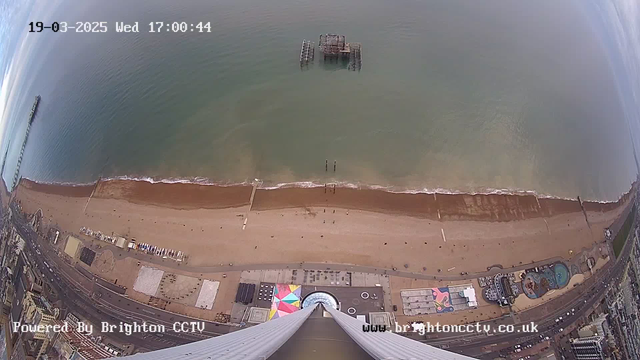 Aerial view of a coastline featuring a sandy beach with a promenade running alongside it. In the background, the ocean is calm with gentle waves lapping at the shore. A distant pier extends into the water, while a partially submerged structure is visible offshore. The beach is mostly empty, with a few people walking, and there are colorful beach umbrellas and structures on the sand. The image is captured from a high perspective looking downwards.