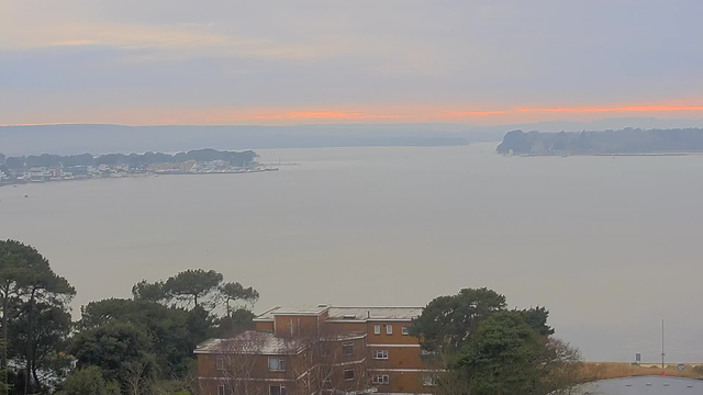 A panoramic view of a calm harbor at dawn. In the foreground, there is a building with multiple windows surrounded by trees. The water is smooth, reflecting the soft pastel colors of the cloudy sky, with a hint of orange on the horizon. In the distance, a small marina is visible with boats and a shoreline lined with buildings and trees. The overall atmosphere is peaceful and serene.