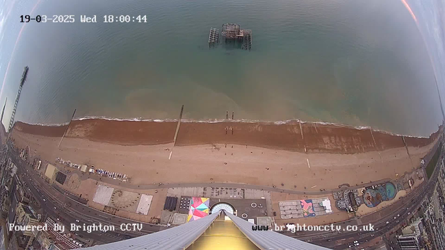 A high-angle view of a sandy beach with several people walking along the shore. In the background is a body of water, transitioning from calm blue to light brown near the beach. A pier extends into the water on the left side, and remnants of an old structure, likely a pier, can be seen partially submerged. There are colorful beach umbrellas and structures near the beach. The image includes a timestamp in the upper left corner, indicating the date and time, and a logo at the bottom for Brighton CCTV.