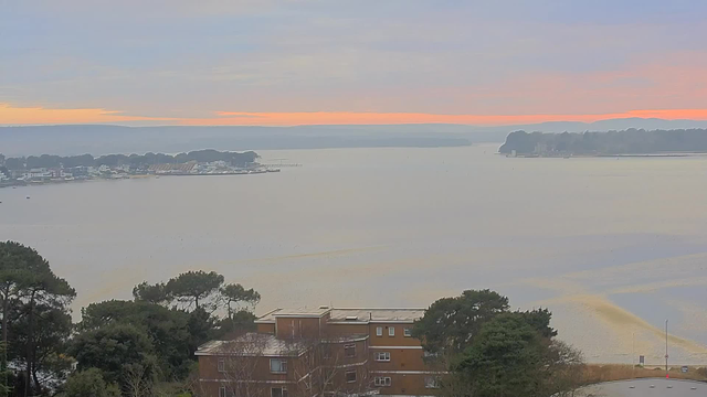 A view of a tranquil body of water with soft ripples, set against a gradient sky transitioning from pale blue to shades of orange and pink near the horizon. In the foreground, there are several low buildings partially obscured by lush green trees, while a distant shoreline is lined with boats and additional structures. The overall scene conveys a serene, early morning atmosphere.