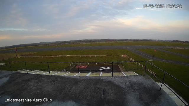 A view from a high vantage point at Leicestershire Aero Club, overlooking a large open field and an airstrip. The foreground features a railing on the building's terrace, with a helipad marked by a large white cross and red lines. The airstrip is visible in the distance, bordered by a wooden fence, and the landscape is mostly flat with patches of grass and sky. The sky above shows soft pastel colors indicating dusk.